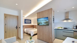 Une chambre d’hôtel moderne avec une porte en bois, un lit, une petite table avec une plante, un tableau et un coin cuisine.
