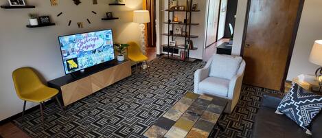 A living room with a TV, yellow chairs, a patterned rug, a grey armchair, and a bookshelf.