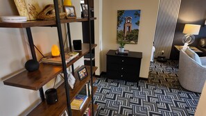 Een hotelkamer met een kamer met een patroon vloer, een zwart dressoir, een houten plank met boeken en een lamp, en een lijst met een afbeelding aan de muur.