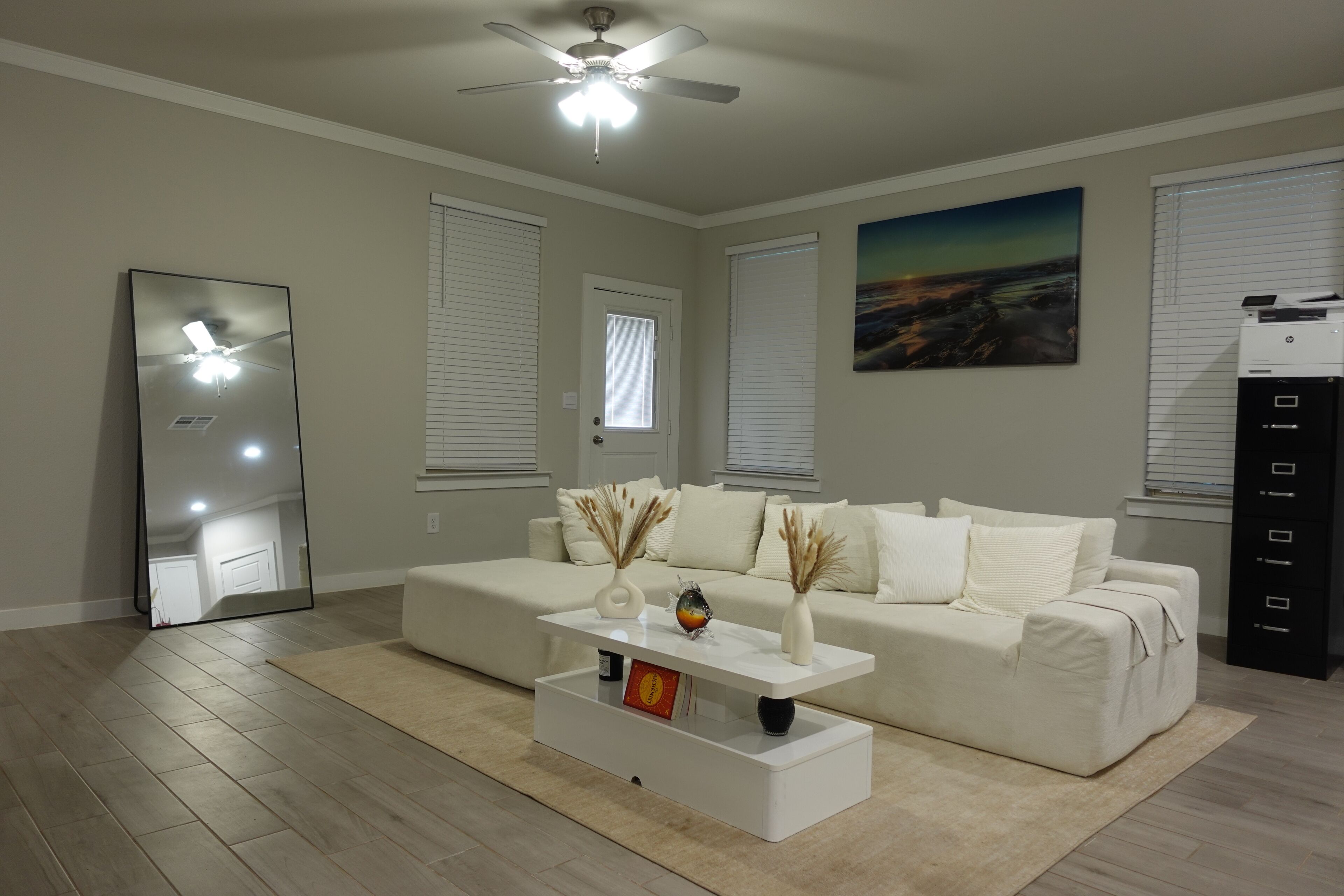 A modern living room with a white sofa, a coffee table, and a ceiling fan. There are two windows with blinds, a painting on the wall, and a printer on the right.