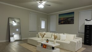 Ein modernes Wohnzimmer mit einem weißen Sofa, einem Couchtisch und einem Deckenventilator. Es gibt zwei Fenster mit Jalousien, ein Gemälde an der Wand und einen Drucker rechts.