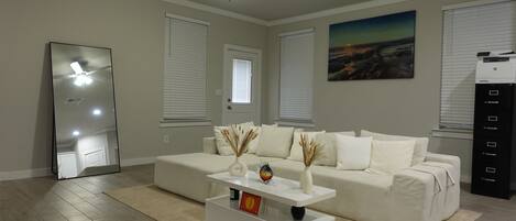 A modern living room with a white sofa, a coffee table, and a ceiling fan. There are two windows with blinds, a painting on the wall, and a printer on the right.