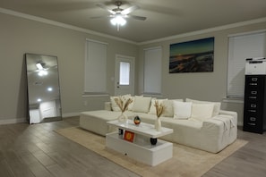 A modern living room with a white sofa, a coffee table, and a ceiling fan. There are two windows with blinds, a painting on the wall, and a printer on the right. - Villa Horizonte Deluxe (Midland)