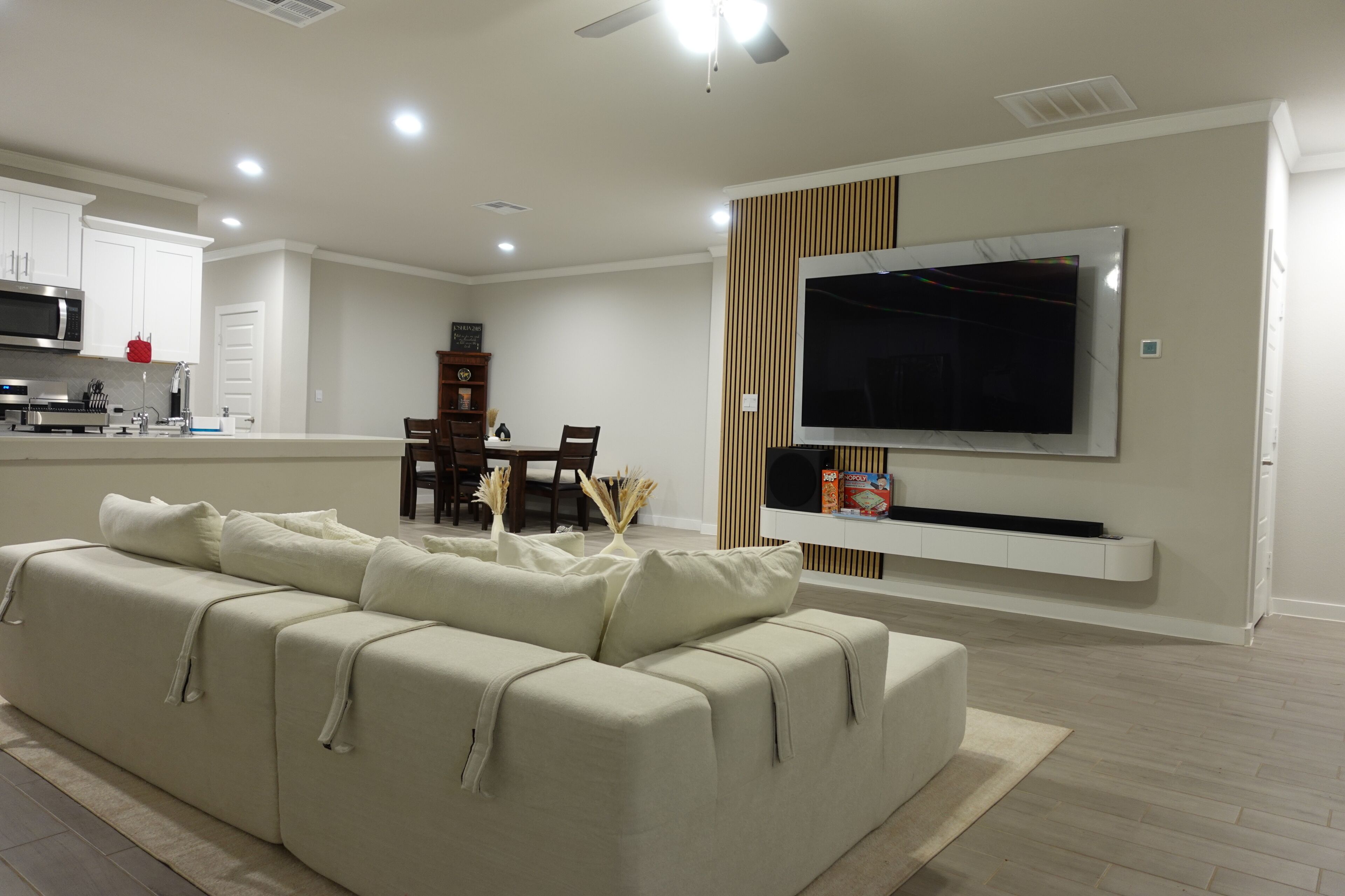 A modern living room with a sofa, a flat-screen TV mounted on the wall, a dining area with chairs and a table, and a kitchen with appliances.