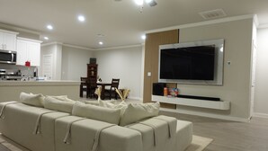 Ein modernes Wohnzimmer mit einer Couch, einem flachbildfernseher an der Wand, einer Essecke mit Stühlen und einem Tisch sowie einer Küche mit Geräten.
