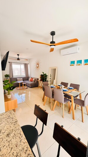 A modern living room with a dining area, a ceiling fan, a flat-screen TV, and a large window with curtains.