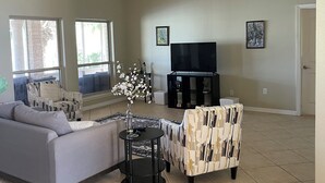 Uma sala de estar ampla com ventilador de teto, um sofá, uma poltrona estampada, uma mesinha com um vaso e uma TV sobre um móvel.