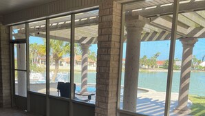 Uma área de estar coberta ao ar livre com móveis de vime, janelas amplas e vista para uma piscina e palmeiras.