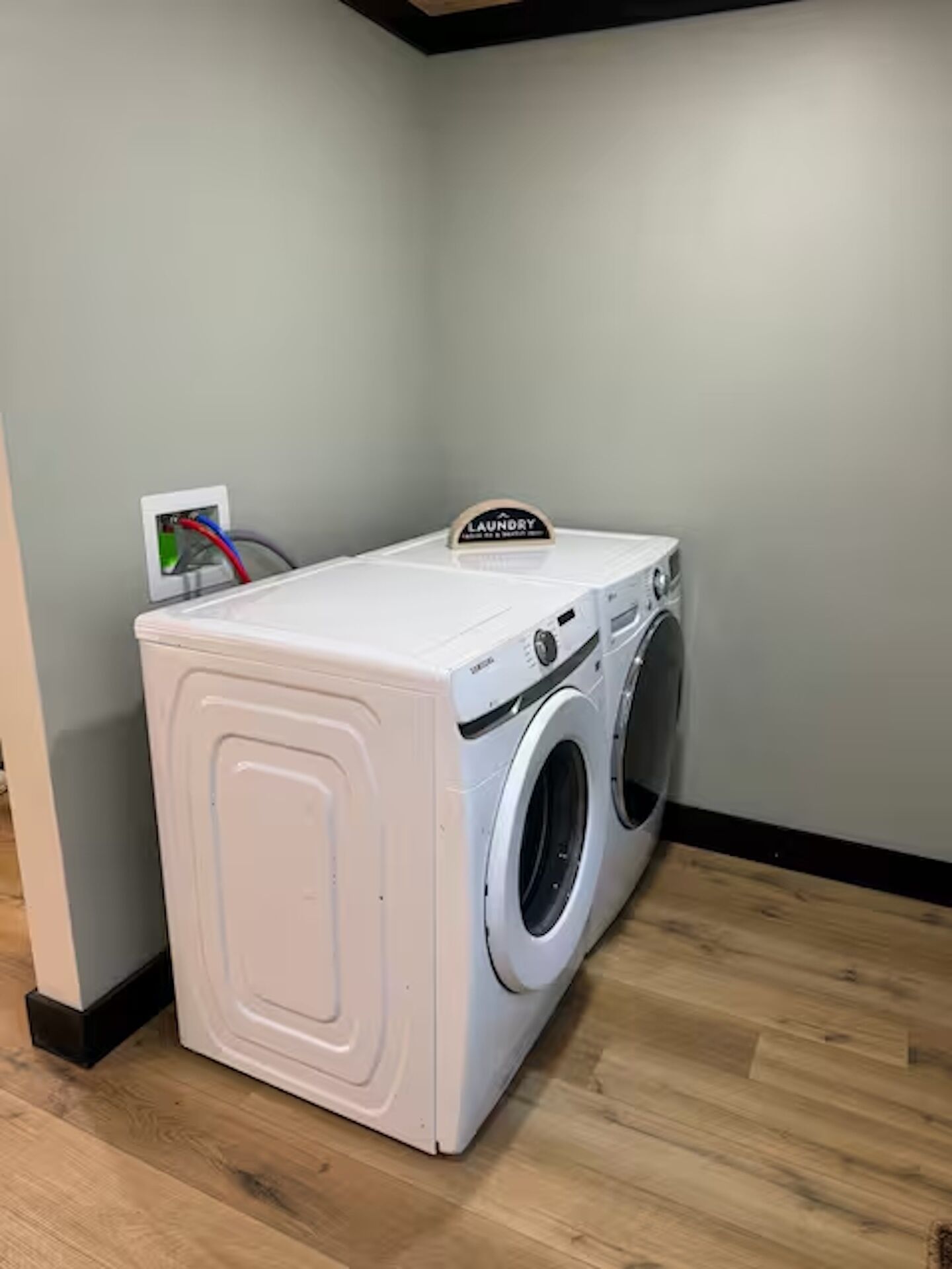 Máquina de lavar roupa de carregamento frontal branca com uma placa 'Lavanderia' em cima, colocada num canto com chão de madeira e parede simples.