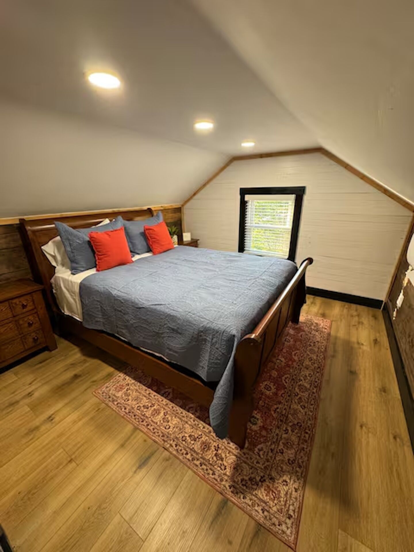 A bedroom with a wooden bed, a nightstand, a window with blinds, and a wooden floor.