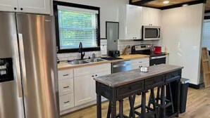 A modern kitchen with stainless steel appliances, white cabinetry, a wooden island with stools, and a window with blinds.