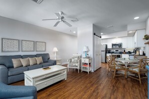 A modern living room with a sofa, coffee table, and dining area with chairs. A kitchen with appliances and cabinets is visible in the background. - Harbour House #301 "Harbour Retreat" (Fort Myers Beach)