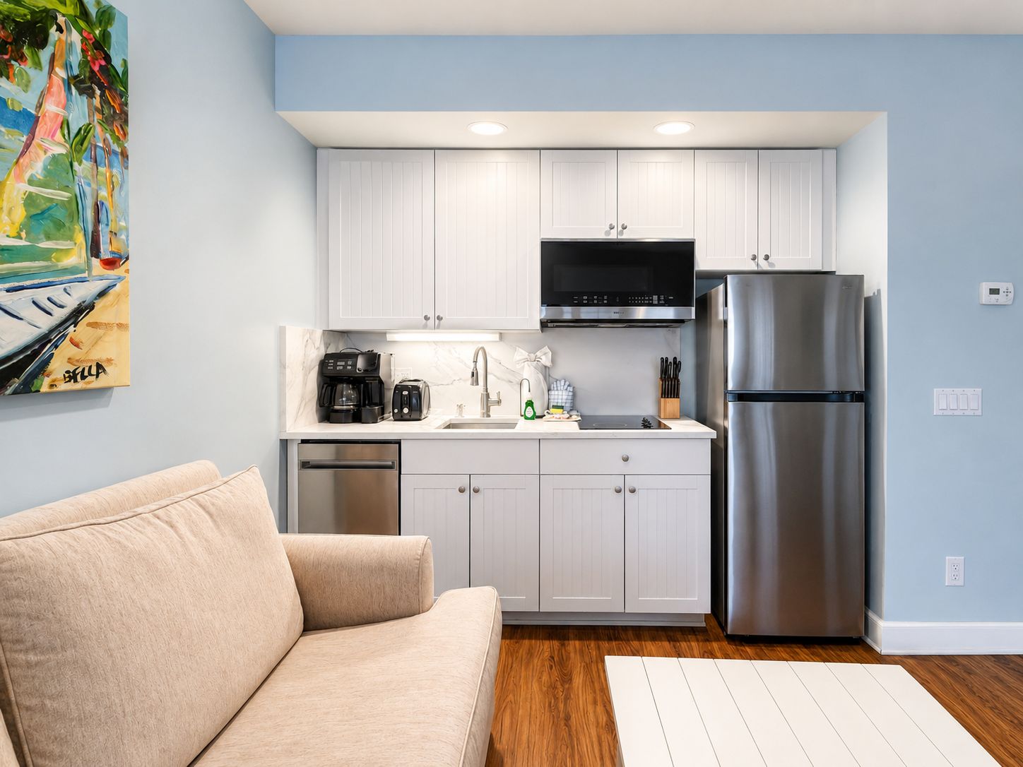 Cocina compacta con armarios blancos, refrigerador de acero inoxidable y cafetera.