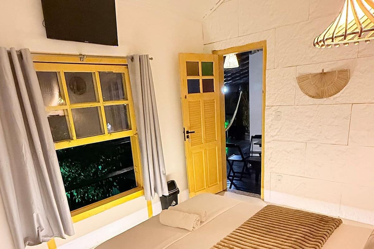 Ein Zimmer mit einem Bett, einem Vorhangfenster, einer Tür, einem an der Wand montierten Fernseher und einer Pendelleuchte.