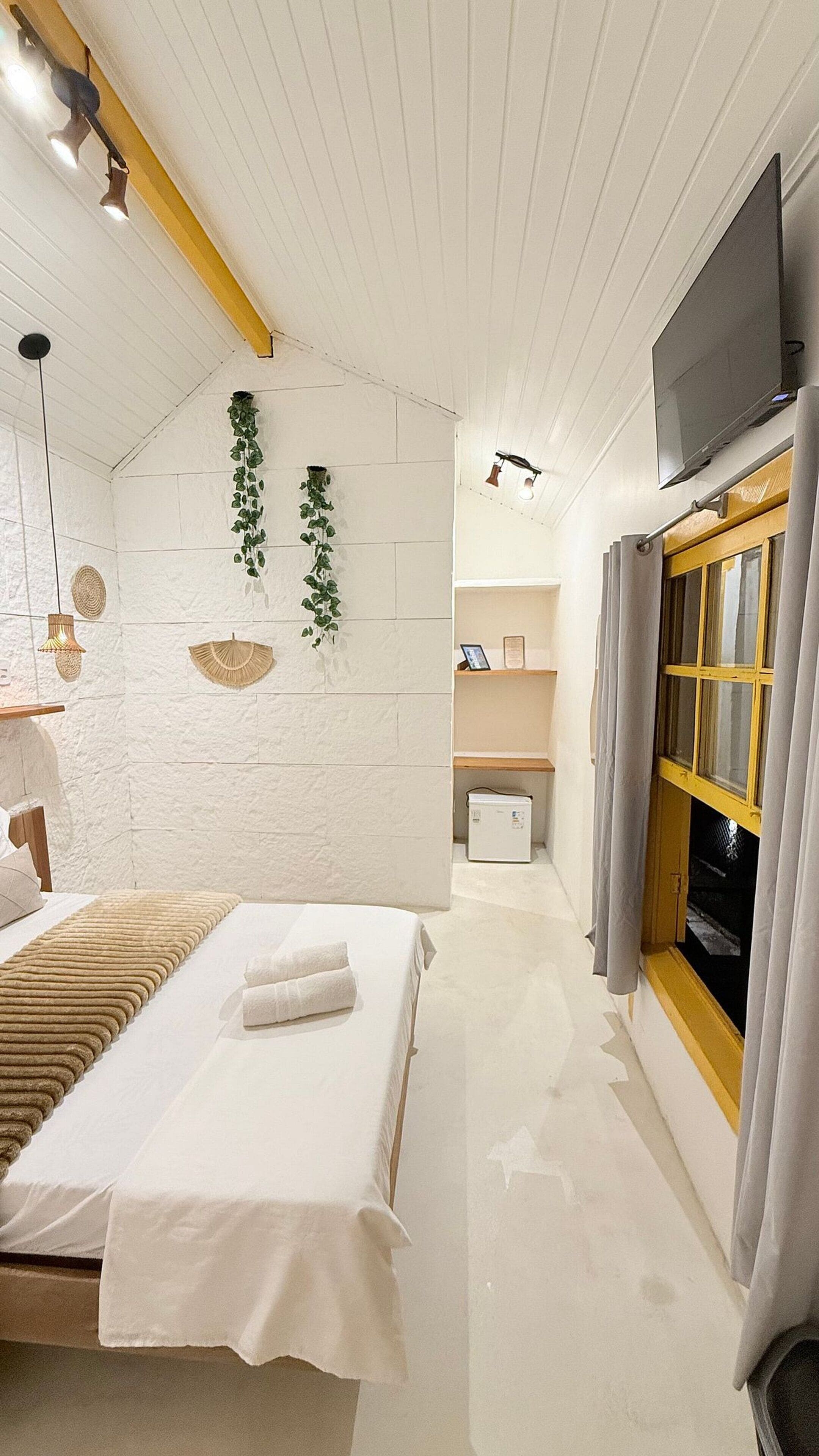 A bedroom with a bed, bedside tables, a TV, a window with curtains, and a plant on the wall.