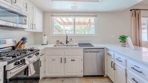 A modern kitchen with white cabinets, a stainless steel stove, a dishwasher, and a window with a view of greenery.