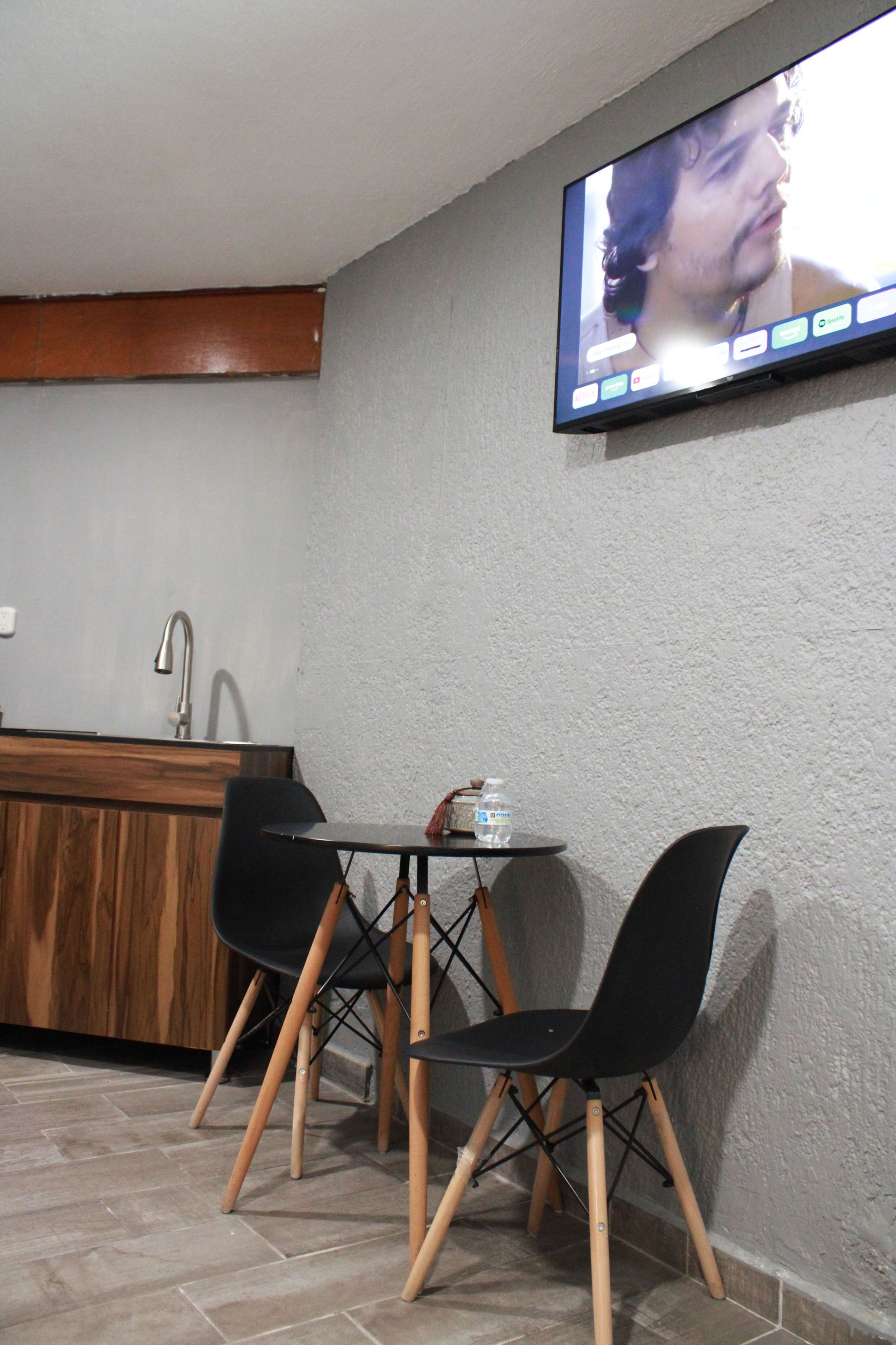 Un petit coin repas avec une table et deux chaises, un téléviseur fixé au mur et, au fond, un espace cuisine.