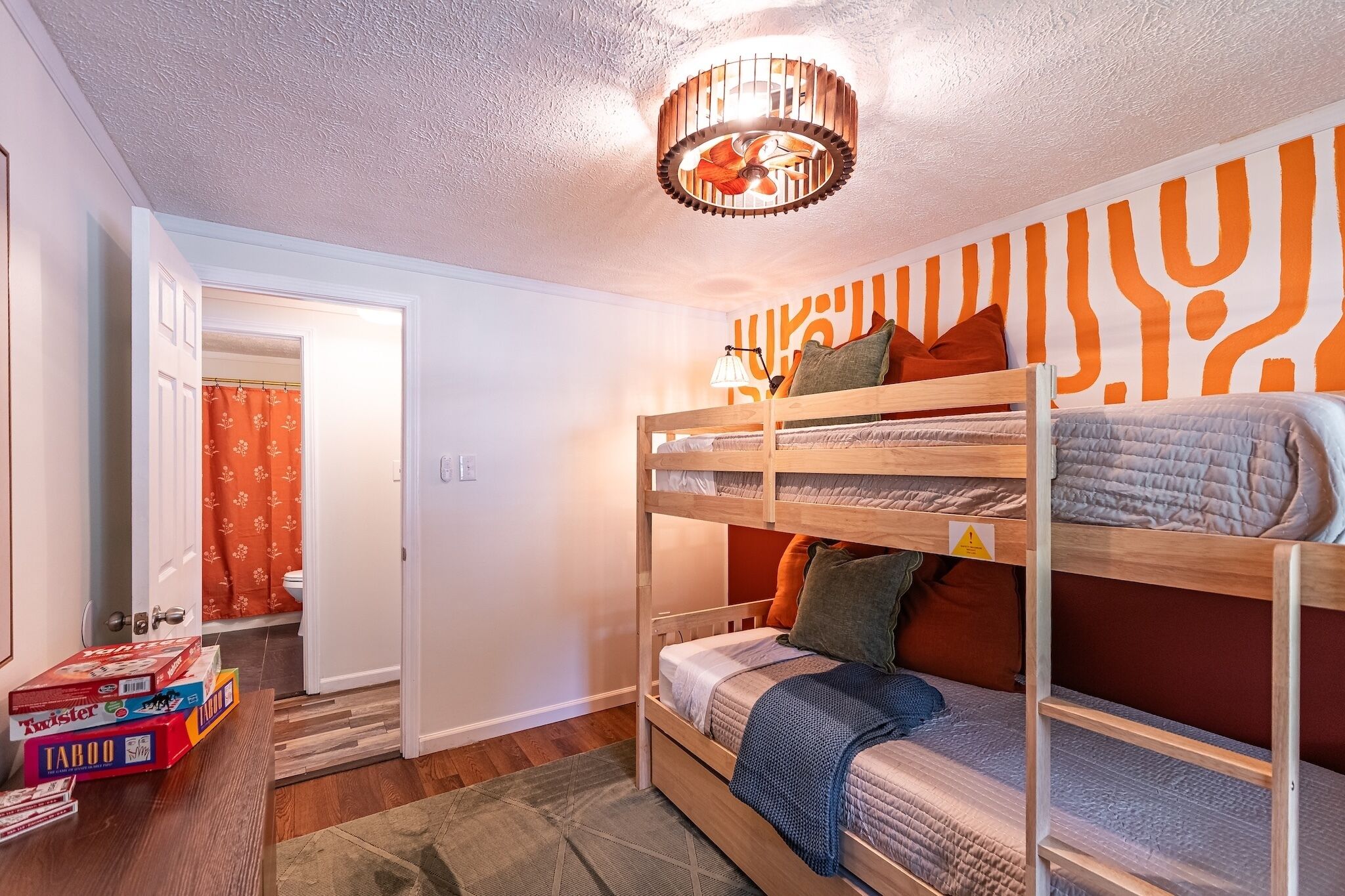 Une chambre avec un lit superposé, un ventilateur de plafond, un mur décoré de motifs et une porte donnant sur une salle de bain.