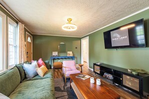 En stue med en grøn sofa, et træsofabord, en pink lænestol, et tv på væggen og en loftsvifte.