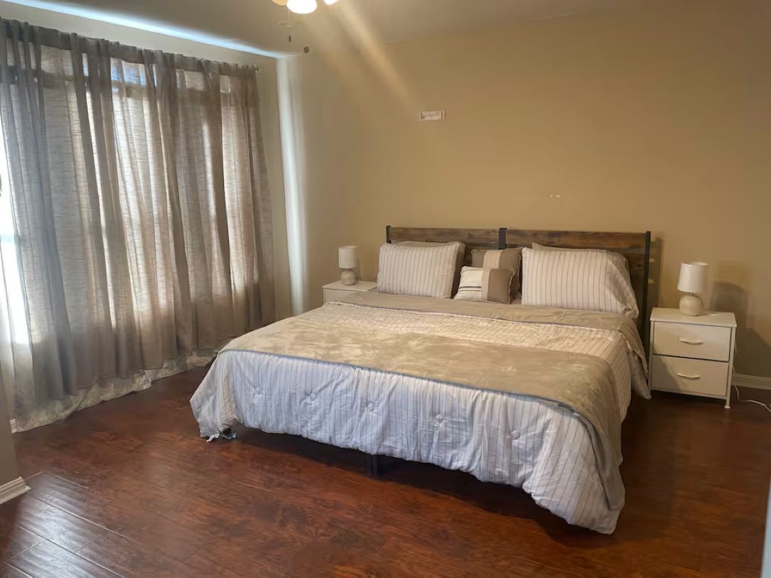 A bedroom with a large bed, a nightstand with a lamp, and a window with curtains.