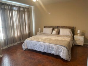 A bedroom with a large bed, a nightstand with a lamp, and a window with curtains. - Lofty Grace Home 2 (Richmond)