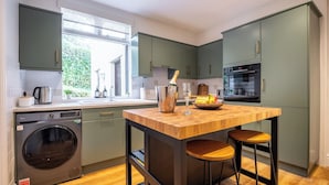 A modern kitchen with a built-in washing machine, a central island with stools, and a view of greenery outside. - Marina View (Windermere)