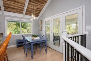 Un coin repas avec une table et des chaises bleues, un banc en bois et une vue sur une terrasse extérieure, accessible par de grandes portes vitrées.