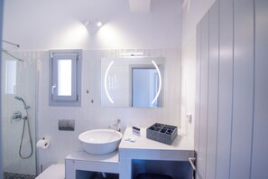 Un baño moderno con lavamanos blanco, cabina de ducha con mampara de vidrio, espejo con iluminación perimetral y una ventana pequeña.