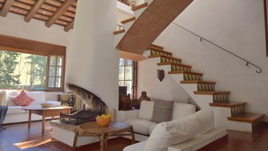 Ampla sala de estar com sofá branco, mesa de vime, lareira e escada de madeira.