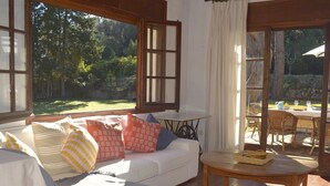 Uma sala de estar acolhedora com um sofá branco, uma mesa pequena e vista para um jardim através de grandes janelas.
