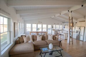 A spacious living area with a large beige sectional sofa, a glass coffee table, and a kitchen in the background. - Modern Coastal Escape/ Ocean Views  4BR/4BA /AC (New Shoreham)