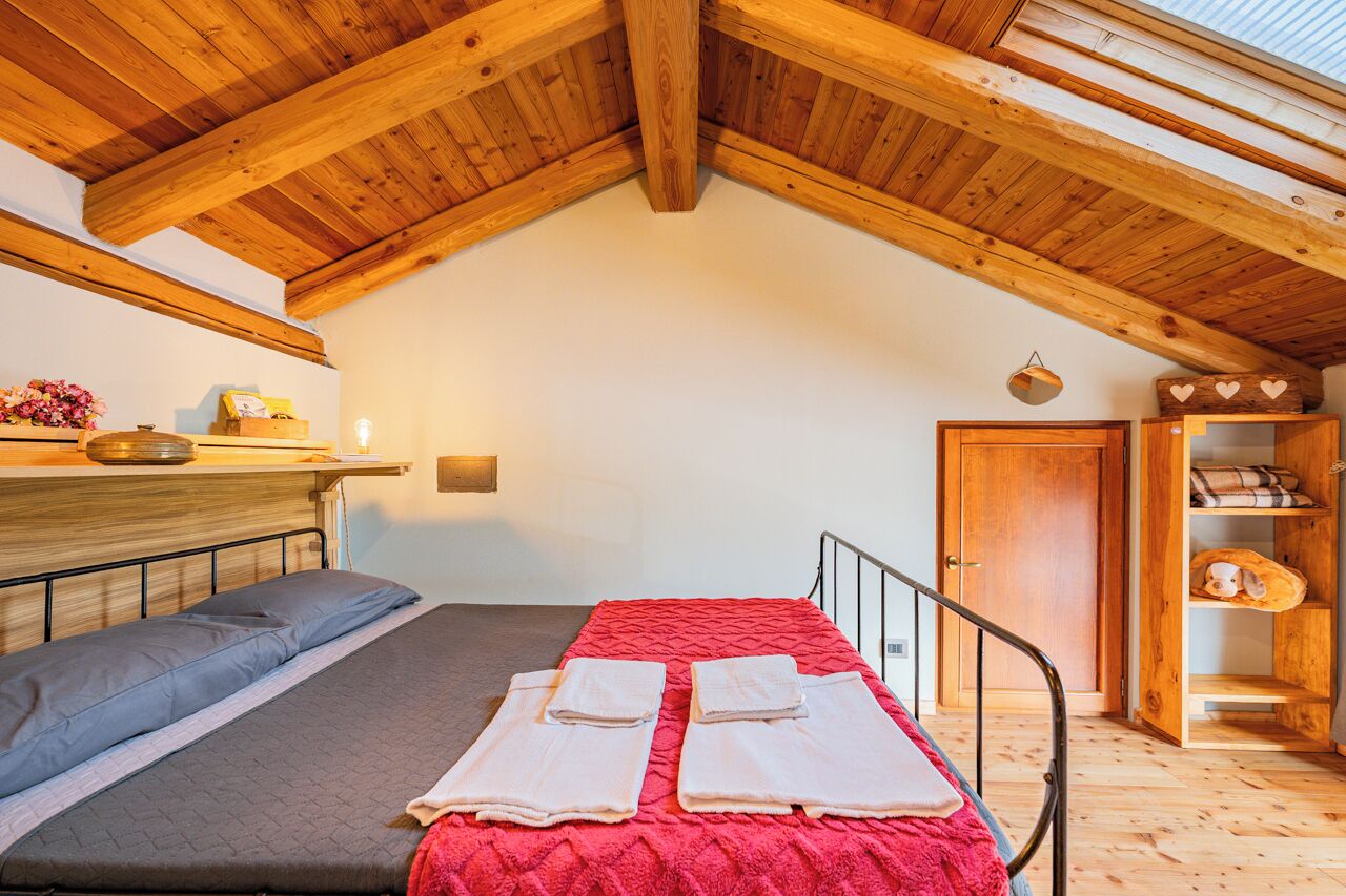 Une chambre mansardée chaleureuse, avec un plafond en bois incliné, un lit recouvert d’une couverture rouge, une étagère en bois ornée de fleurs et une armoire en bois.