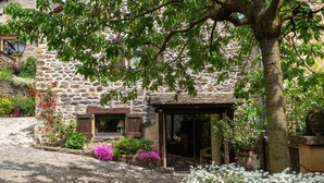A stone building with a covered entrance, a window with shutters, and a garden with flowers and a tree. - La grande du Bruel - ESCLANEDES (ESCLANEDES)