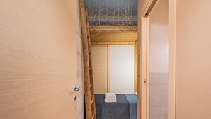 Ein schmaler Flur, der zu einem Raum mit einem Bett, einer Holzleiter und einer Tür führt.