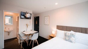 Et hotelværelse med seng, natbord, lampe, fjernsyn, skrivebord med stole og et håndvask.