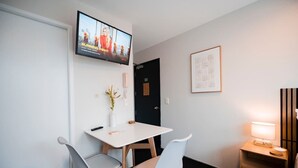 Et hotelværelse med fjernsyn monteret på væggen, et skrivebord med to stole, et sengebord med lampe og et billede i ramme på væggen.