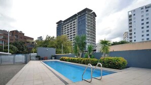 En swimmingpool med en metalstige, omgivet af en fliseterrasse, med bygninger i baggrunden.