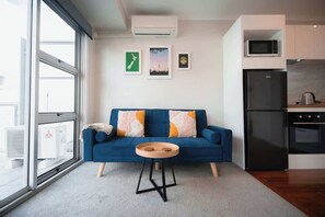 A modern living room with a blue sofa, a small wooden side table, and a kitchen area with a black refrigerator and microwave. - Princes Wharf Harbour View Studio (Auckland Central)