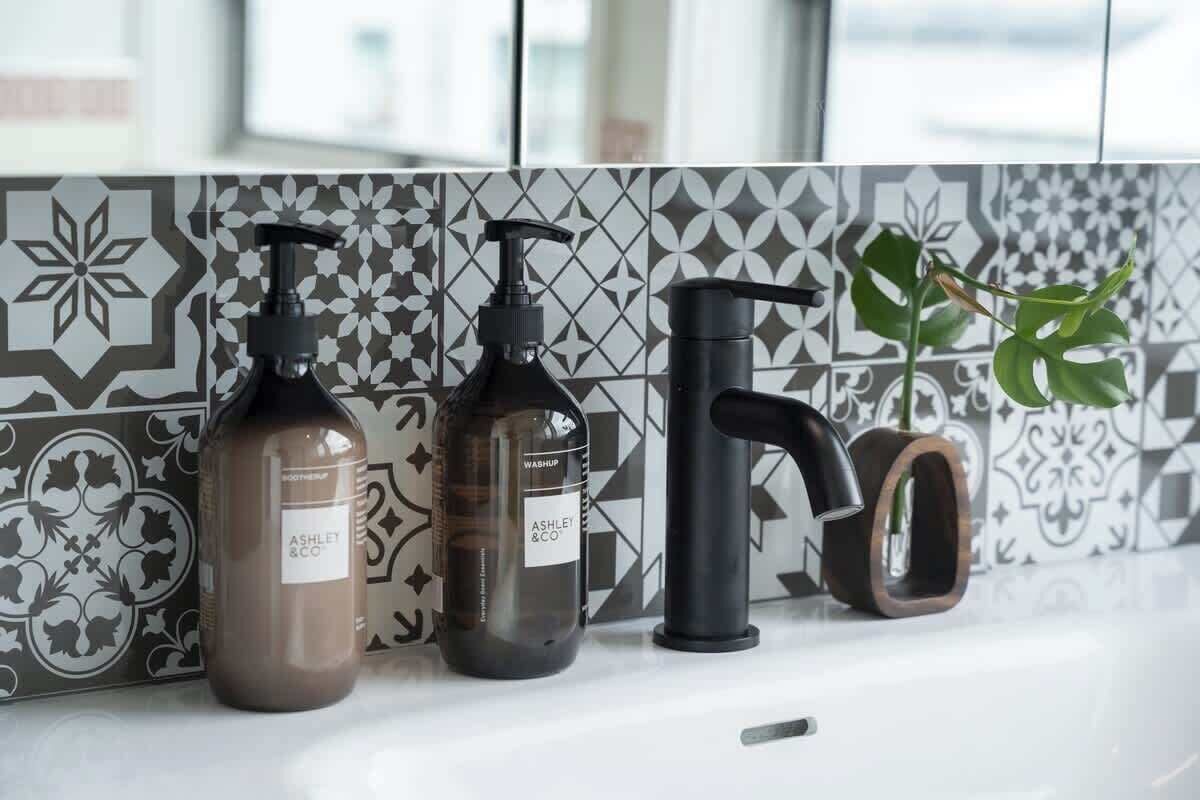 A bathroom with patterned tiles, two bottles of liquid soap, a black faucet, and a potted plant.