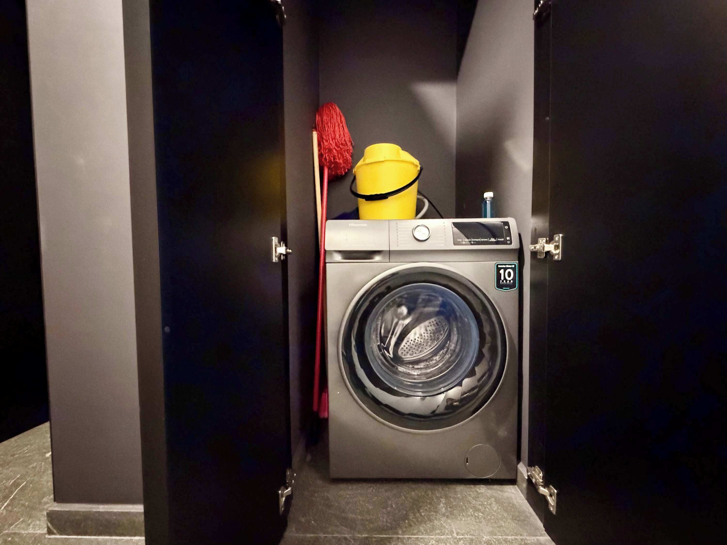 Eine kompakte, moderne Waschmaschine in einem kleinen, geschlossenen Raum, zusammen mit einem Mop und einem gelben Eimer.