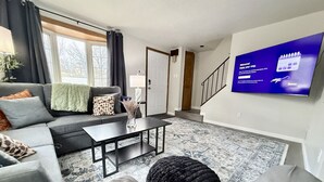 Una sala de estar con un sofá, una mesa de centro, un reposapiés y un televisor de pantalla plana montado en la pared.