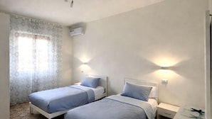 Ein Zimmer mit zwei Betten, einem Fenster mit Vorhängen, einer Klimaanlage und einem Nachttisch mit Lampe.