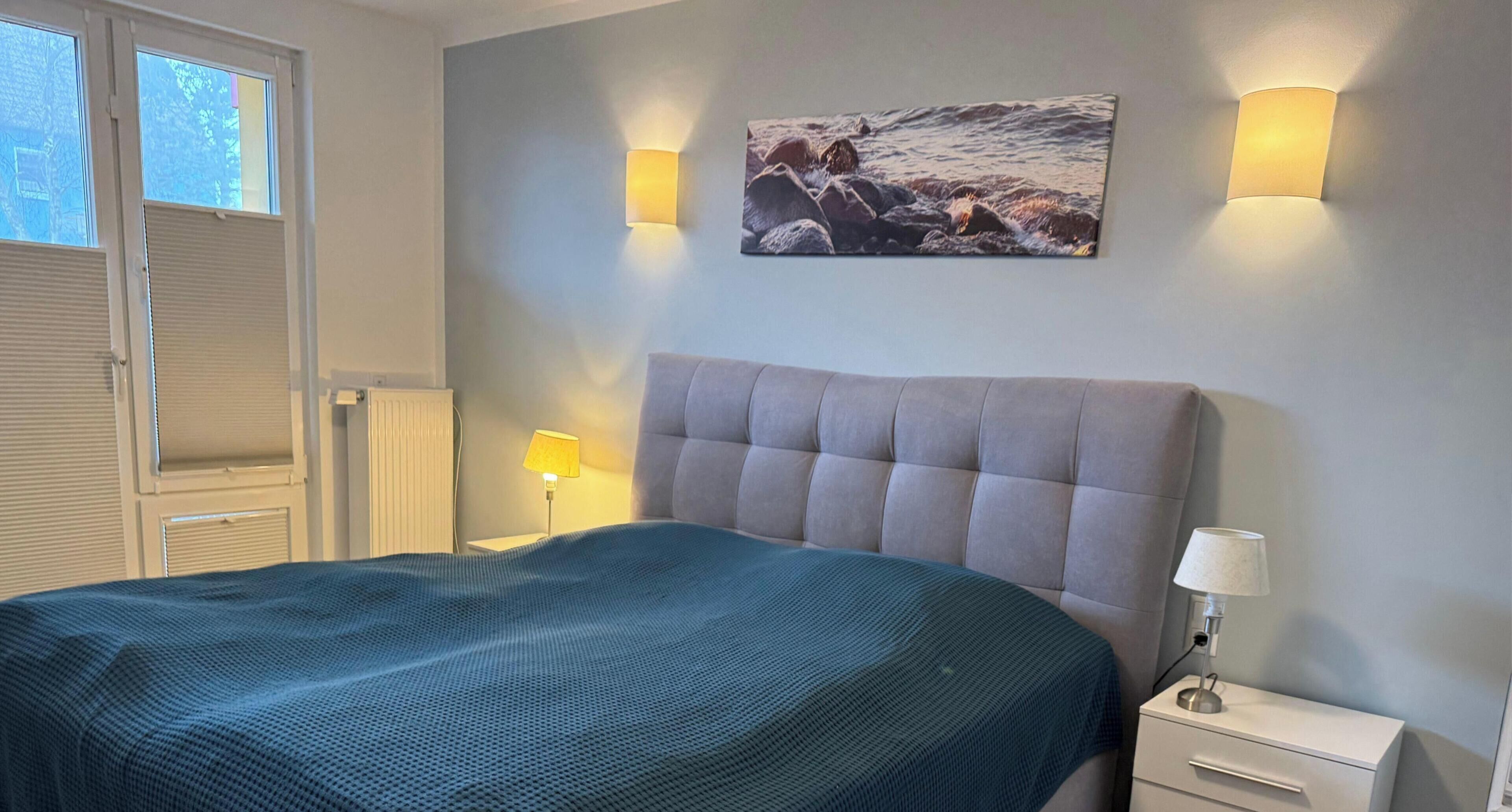 A bedroom with a bed, a nightstand, a lamp, a wall-mounted picture, and a window with blinds.