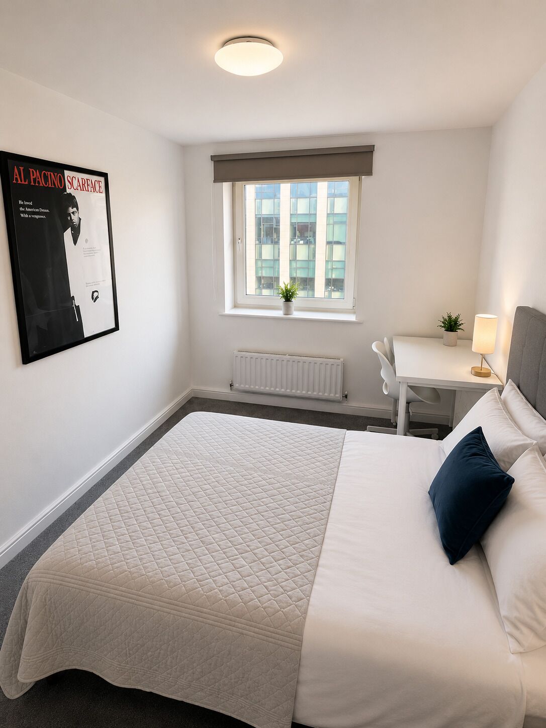 Een hotelkamer met een bed, bureau, stoel, radiator, raam en een poster aan de muur.