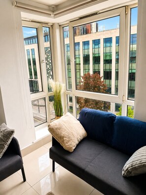 A modern living room with a sofa, a small table with a plant, and large windows offering a view of buildings and a street. - Spacious 3-Bedroom Flat in Central Leeds (Leeds)