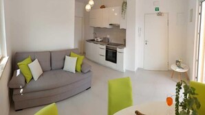 Un salon moderne avec un canapé gris, une petite table à manger avec des chaises, un coin cuisine avec des meubles blancs et une porte portant un panneau indiquant la sortie.