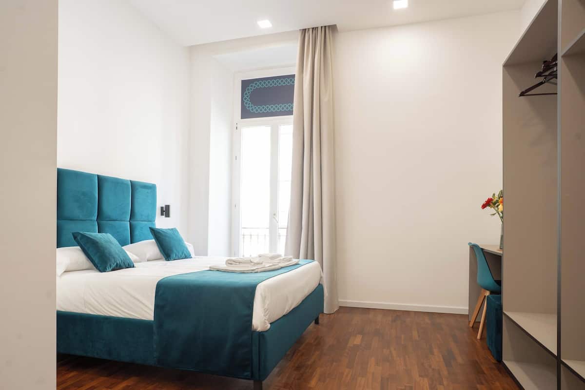 A modern bedroom with a teal upholstered bed, white bedding, a wooden floor, a small desk with a chair, and a window with curtains.