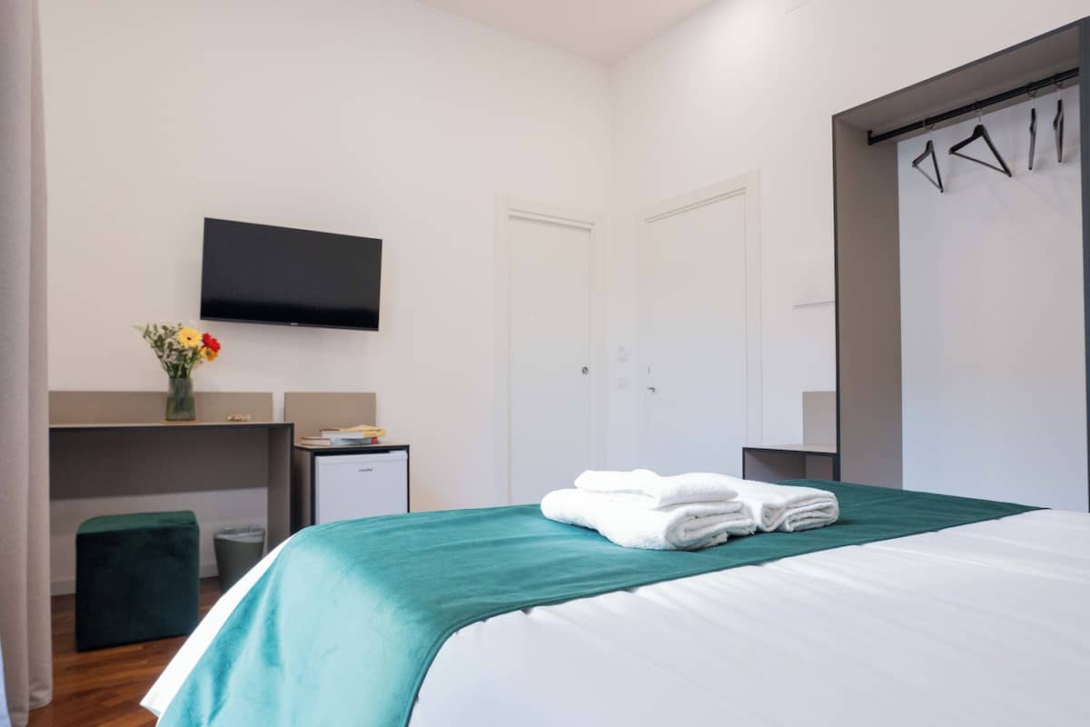 A hotel room with a bed, a flat-screen TV, a desk with a computer, a small trash bin, and a wardrobe with hangers.