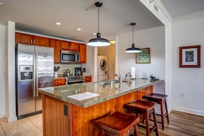 A modern kitchen with wooden cabinets, stainless steel appliances, granite countertops, and bar stools. - Honua Kai 2 Free Rental Cars KBM Resorts Spacious Floorplan 2 Units, 3 Bedrooms HK Ml-2717 (Kaanapali)