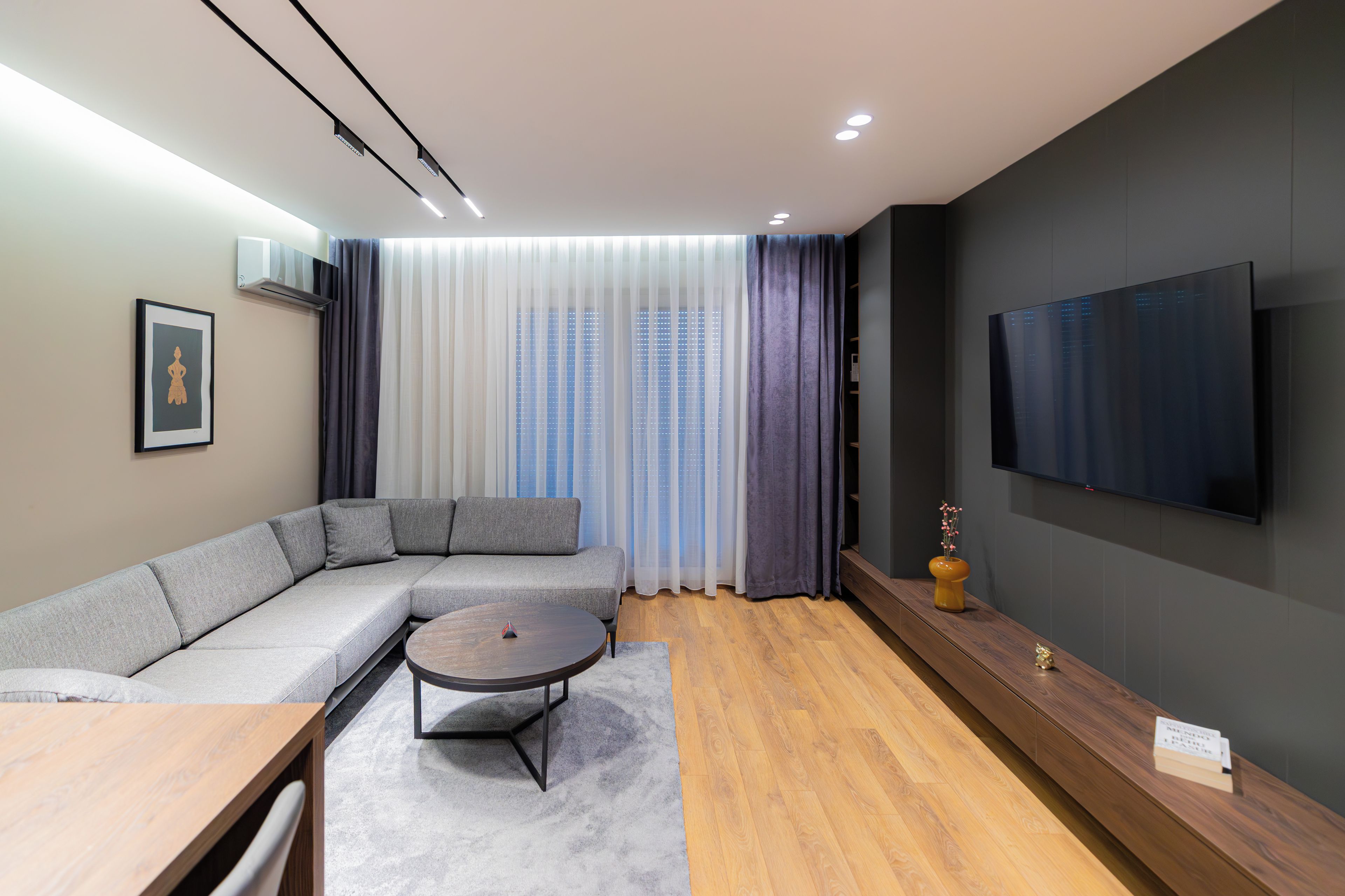 A modern living room with a sectional sofa, a coffee table, a flat-screen TV, and a large window with curtains.